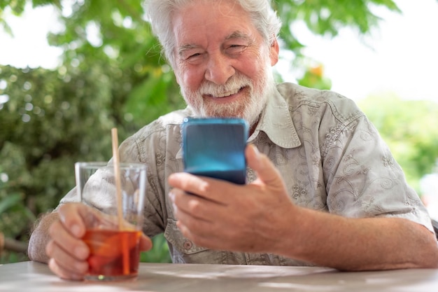 Un anciano feliz sentado al aire libre en una mesa de café sosteniendo un vaso y mirando el teléfono