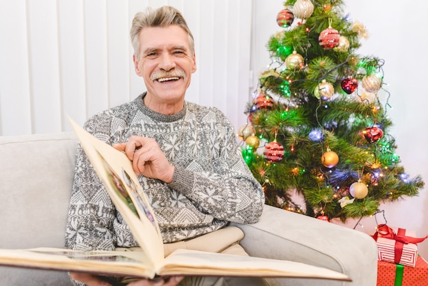 El anciano feliz sostiene un álbum de fotos cerca del árbol de navidad