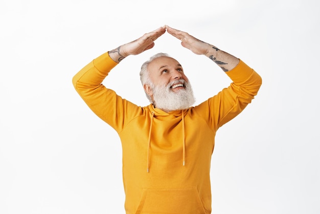 Un anciano feliz con tatuajes mirando sus manos en la azotea, un anciano que hace un gesto de casa en el techo y una sonrisa alegre de pie contra un fondo blanco con una capucha naranja