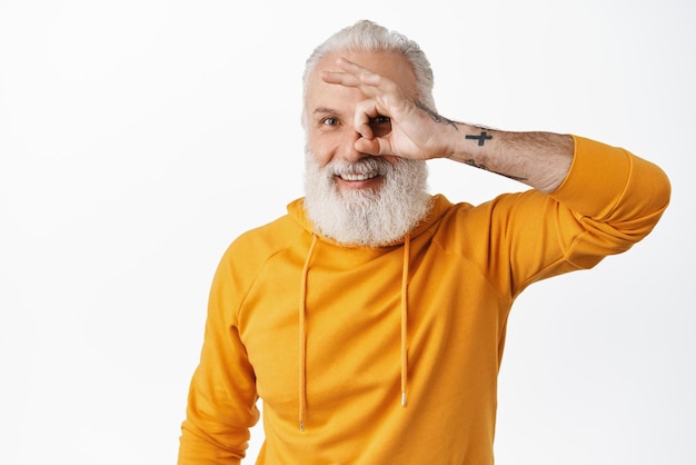 Un anciano feliz con tatuajes que muestra el signo de estar bien mirando a través de los dedos bien y sonriendo complacido satisfecho elogio algo bueno decir sí o bien de pie sobre fondo blanco