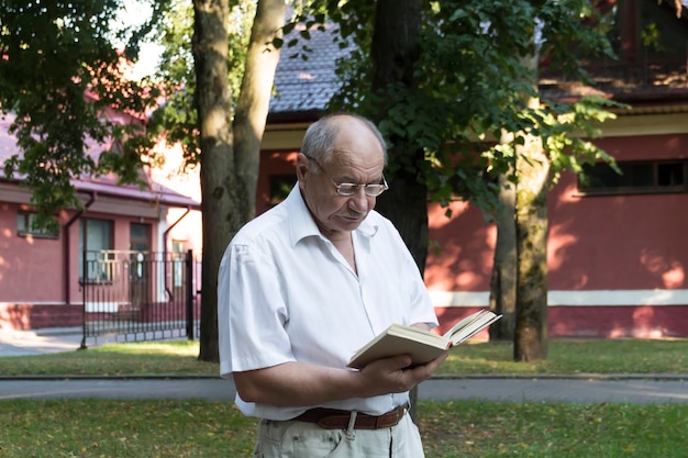 Un anciano jubilado camina solo en el parque Un anciano calvo con anteojos se para contra el fondo de un edificio rojo y lee un libro