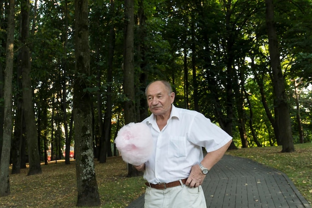 Un anciano jubilado con una camisa blanca camina con un enorme algodón de azúcar rosa en el camino del parque