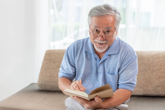 Anciano mayor asiático con gafas leyendo un libro de novelas, sonriendo se siente feliz en el sofá en casa - concepto de personas mayores mayores de estilo de vida