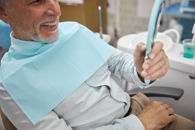 un anciano mirando en el espejo y sonriendo mientras descansa en el sillón dental