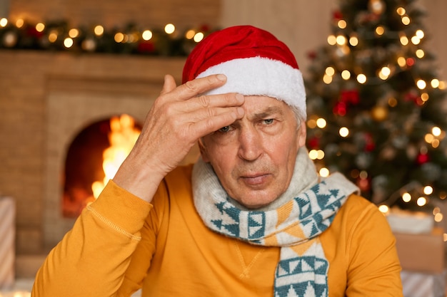 Anciano molesto infeliz cansado que está enfermo en la víspera de Navidad, que sufre de dolor de cabeza, que mantiene la mano en la frente, que lleva camisa amarilla y gorro de Papá Noel, con expresión molesta.