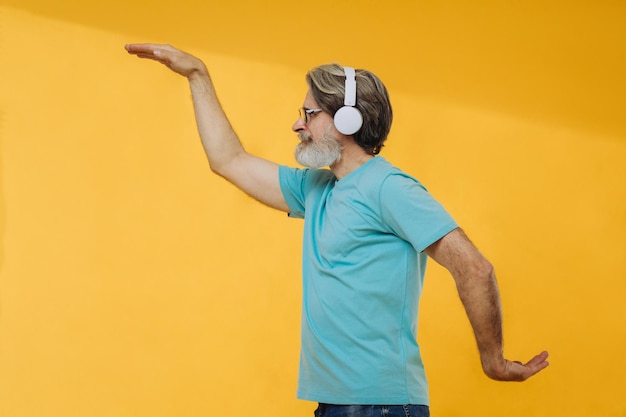 Un anciano optimista de pelo gris escucha música baila y lleva auriculares con gafas aisladas en un fondo amarillo