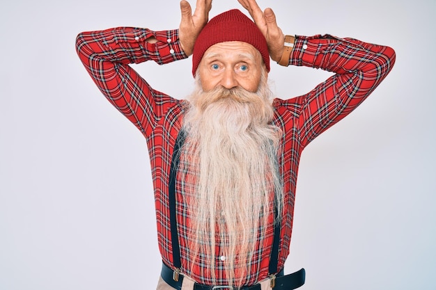 Un anciano de pelo gris y barba larga con aspecto de hipster con gorro de lana haciendo gestos de orejas de conejo con las palmas de las manos con aspecto cínico y escéptico. concepto de conejo de pascua.