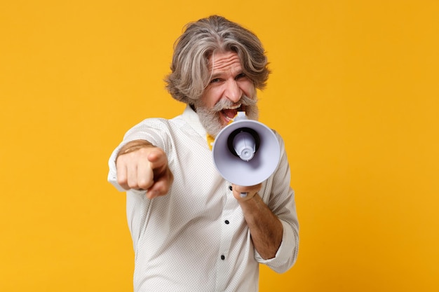 Anciano de pelo gris con bigote barbudo en camisa blanca pajarita aislado sobre fondo amarillo en el estudio. Concepto de estilo de vida de las personas. Simulacros de espacio de copia. Gritar en megáfono señalar con el dedo índice a la cámara.