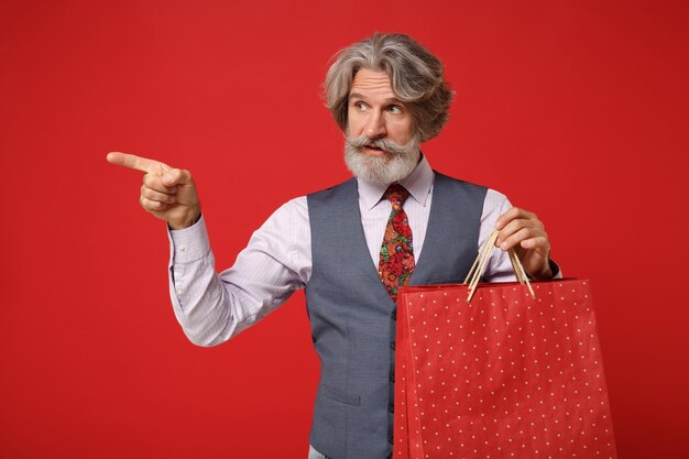 Anciano de pelo gris bigote barbudo hombre en camisa chaleco corbata aislado sobre fondo rojo. Concepto de estilo de vida de las personas. Simulacros de espacio de copia. Sostenga la bolsa del paquete con la compra después de comprar, señale con el dedo a un lado.