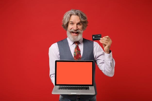 Anciano de pelo gris bigote barbudo hombre en camisa chaleco corbata posando aislado sobre fondo rojo. Concepto de estilo de vida de las personas. Simulacros de espacio de copia. Sostenga una computadora portátil con una tarjeta de crédito de pantalla vacía en blanco.