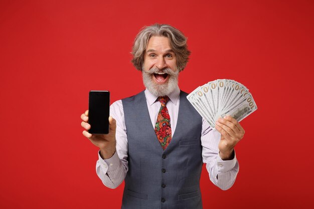Anciano de pelo gris bigote barbudo hombre en camisa chaleco corbata posando aislado sobre fondo rojo. Concepto de estilo de vida de las personas. Simulacros de espacio de copia. Sostenga el teléfono móvil con un ventilador de pantalla vacío en blanco de dinero en efectivo.