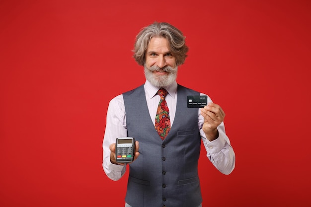 Anciano de pelo gris bigote barbudo hombre en camisa clásica chaleco corbata aislado sobre fondo rojo. Concepto de estilo de vida de las personas. Simulacros de espacio de copia. Sosteniendo terminal de pago de banco inalámbrico moderno, tarjeta de crédito.