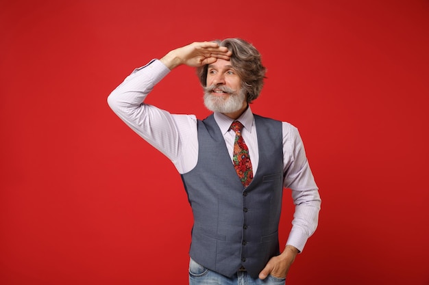 Anciano de pelo gris bigote barbudo hombre en camisa clásica chaleco corbata colorida aislado sobre fondo rojo. Concepto de estilo de vida de las personas. Simulacros de espacio de copia. Mantenga la mano en la frente mirando a lo lejos.