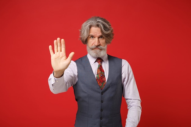 Anciano de pelo gris bigote hombre barbudo en camisa clásica chaleco corbata colorida aislado sobre fondo rojo, retrato de estudio. Concepto de estilo de vida de las personas. Simulacros de espacio de copia. Mostrando gesto de parada con la palma.