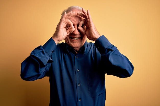 Un anciano de pelo gris con un pantalones azul casual sobre un fondo amarillo haciendo un gesto correcto como binoculares sacando la lengua de los ojos mirando a través de los dedos Expresión loca