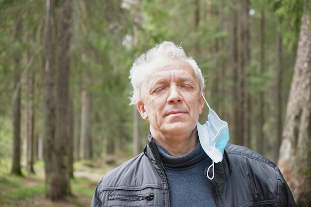 el anciano con el pelo gris se quita una máscara protectora médica y respira profundamente en el bosque