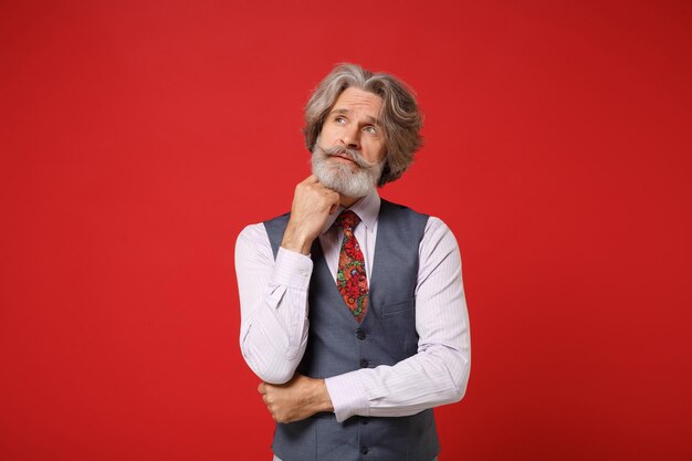 Un anciano pensativo con barba de bigote canoso con camisa clásica, chaleco, corbata colorida, posando aislado en un fondo rojo en el estudio. Concepto de estilo de vida de las personas. Simulacros de espacio de copia. Coloque la mano apoyada en la barbilla.