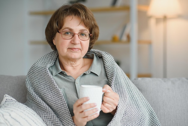 Anciano pensionista caucásico envuelto en tela escocesa gris y bebiendo té caliente en la sala de estar