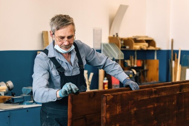 Un anciano pinta tablas de madera con barniz oscuro en un taller de carpintería Un jubilado con una máscara médica