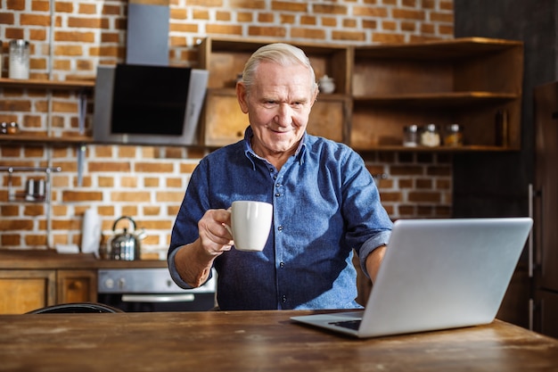 Anciano positivo sonriendo mientras usa la computadora portátil y bebe té en la cocina