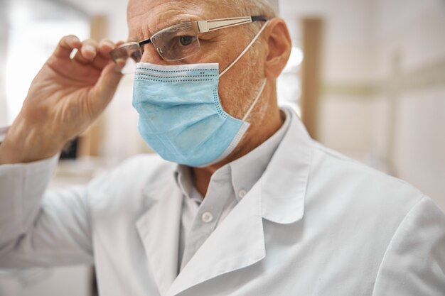 Anciano profesional trabajador de la salud tocando sus gafas y mirando serio en el trabajo