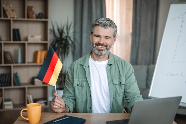 Un anciano profesor europeo sonriente con barba sosteniendo la bandera de alemania cerca de la pizarra en la habitación