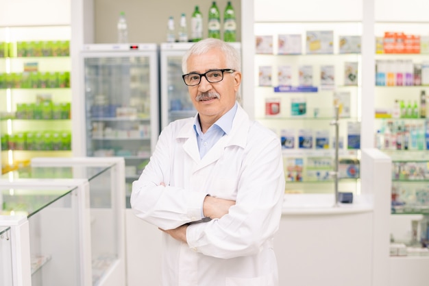 Anciano propietario exitoso de una gran farmacia contemporánea cruzando los brazos por el pecho mientras está de pie contra la pantalla con nueva medicina