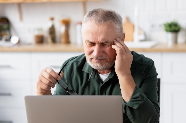 Anciano que sufre de dolor de cabeza severo mientras trabaja en la computadora portátil