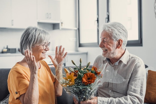 Anciano regalando flores a su esposa sentada en el sofá de casa para el día de San Valentín Pensionistas disfrutando juntos de una sorpresa Enamorados divirtiéndose