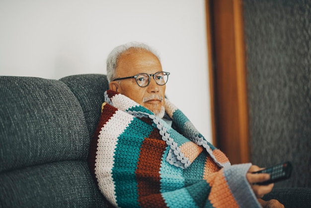 Un anciano sentado en el sofá en casa con una cubierta de lana colorida para síntomas de baja temperatura y gripe fría Concepto de crisis de gas energético Ahorro de calor interior Concepto de personas en invierno