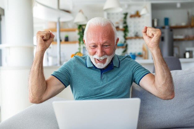 Anciano sentado en el sofá mirando la pantalla de la computadora portátil gritar con alegría se siente emocionado feliz celebrando la victoria de la lotería momento afortunado obtuvo oportunidad en línea ventas y descuentos concepto de comercio electrónico