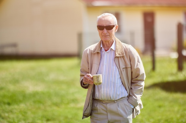 Un anciano serio de 75 años parado afuera frente a su casa con una taza de bebida caliente en la mano y gafas de sol en la cara