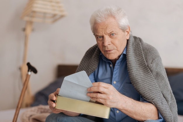 Anciano serio pensativo sentado en la cama y tomando una carta mientras sostiene el buzón