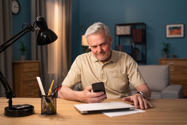 Un anciano se sienta en la mesa de su sala de estar con una lámpara portátil y un teléfono por la noche