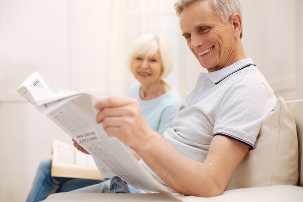 Anciano sonriente analítico leyendo un periódico nuevo y mirando encantado mientras su señora le pregunta por su opinión