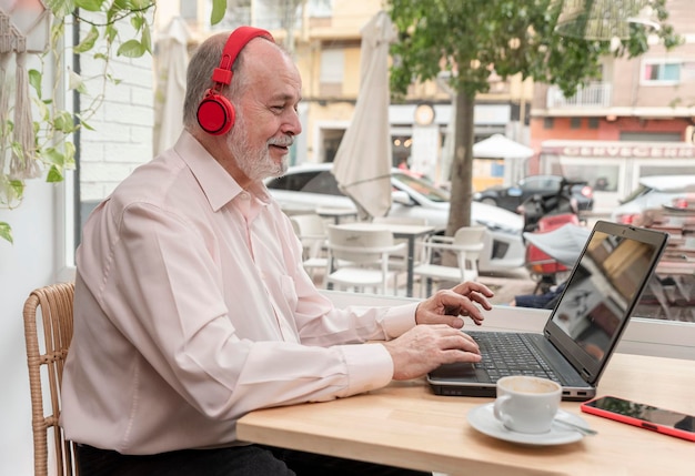 Un anciano sonriente con audífonos mirando la pantalla de una computadora portátil viendo un seminario web o tomando clases en línea sentado en una cafetería con su taza de café a su lado