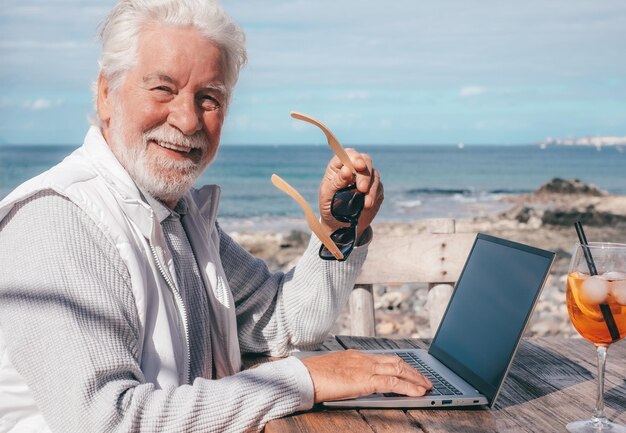 Un anciano sonriente con barba trabaja en una laptop al aire libre en la playa en un día soleado Concepto de libertad