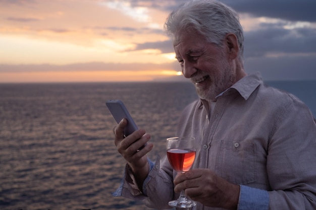 Anciano sonriente con una copa de vino en la mano de pie frente al mar mirando el teléfono móvil mientras el sol se pone en el mar Paz soledad relajación