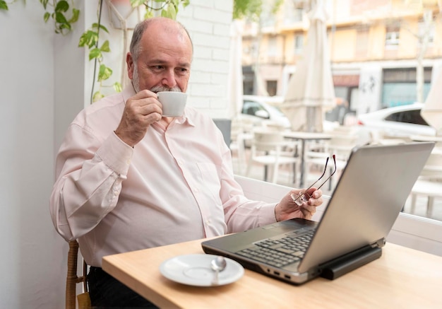 Un anciano sonriente dentro de una cafetería sosteniendo sus anteojos mirando correos electrónicos en su computadora portátil mientras bebe su café