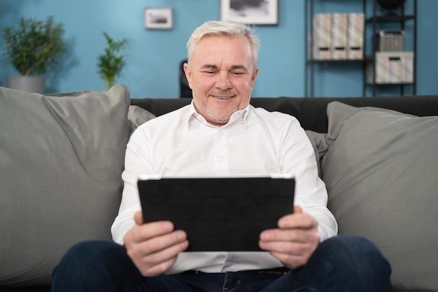 Un anciano sonriente de mediana edad que se relaja sosteniendo una tableta digital leyendo un libro electrónico sentado en un sofá en casa