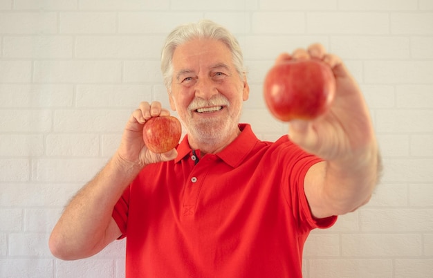 Un anciano sonriente de rojo aislado sobre un fondo blanco sosteniendo dos manzanas rojas en sus manos mirando el concepto de alimentación saludable de la cámara