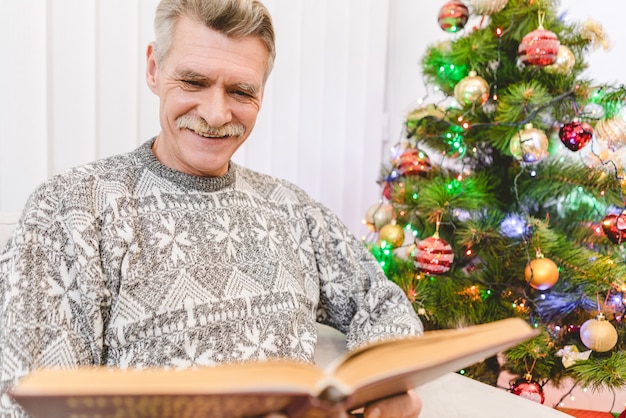 El anciano sostiene un libro cerca del árbol de navidad.