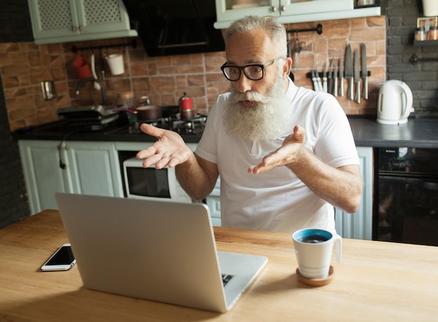 Anciano trabajando en equipo portátil, sonriendo, mirando la pantalla, bebiendo té.
