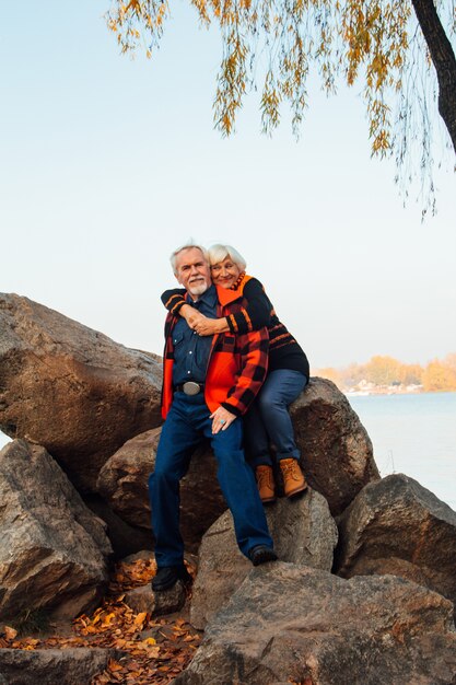 Ancianos alegres, una mujer y un hombre están sentados en las piedras y abrazándose en el lago