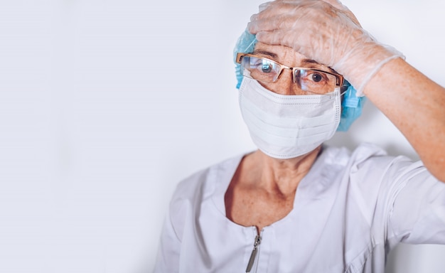 Ancianos cansados cansados mujer madura médico o enfermera sosteniendo la cabeza en una bata blanca médica, guantes, mascarilla con equipo de protección personal. Asistencia sanitaria y medicina. Covid-19 crisis pandémica