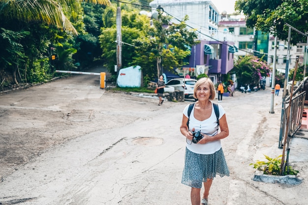 Ancianos mayores mochileros asiáticos viajando mujer madura turista caminando disfrutando de tomar fotos en la calle local de Sanya al aire libre. Viajando por Asia, concepto de estilo de vida activo. Descubriendo Hainan, China