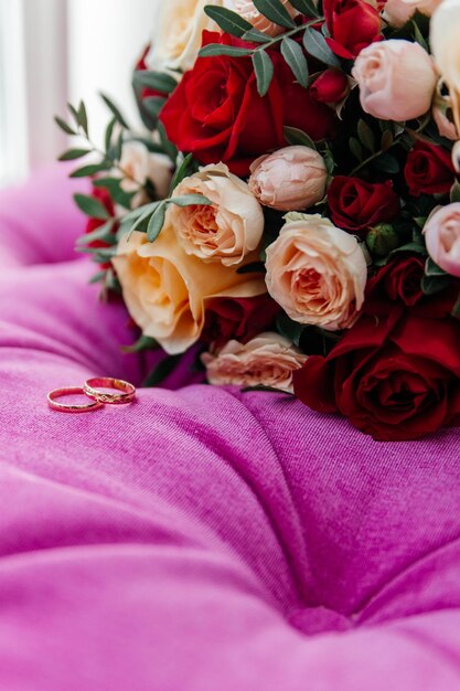 Anéis de casamento dourados ao lado do buquê de rosas vermelhas e amarelas da noiva sobre uma cadeira rosa