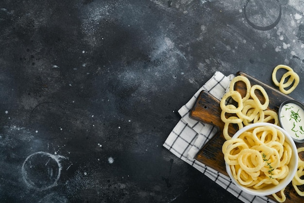 Anéis de cebola Fast food Chips de cebola crocantes em tigela preta cerâmica com molho de creme azedo e cebolas em suporte de madeira no antigo fundo de concreto preto Tradição americana Hot BBQ Vista superior