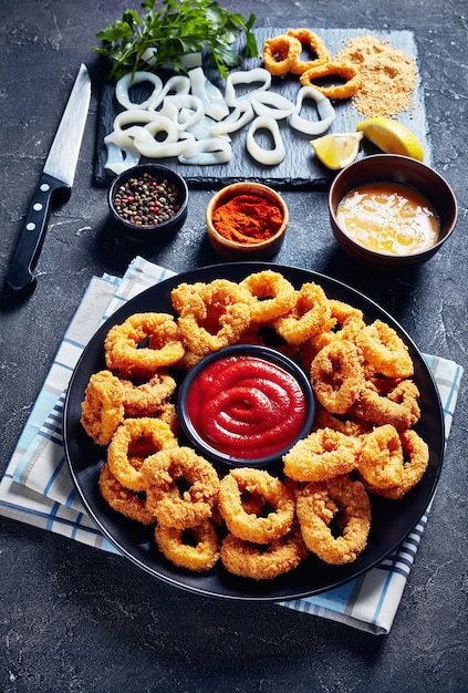 Anéis de lula crocantes, anéis de lula empanados fritos e fritos servidos com molho de tomate em um prato preto sobre uma mesa de concreto com ingredientes, vista vertical de cima