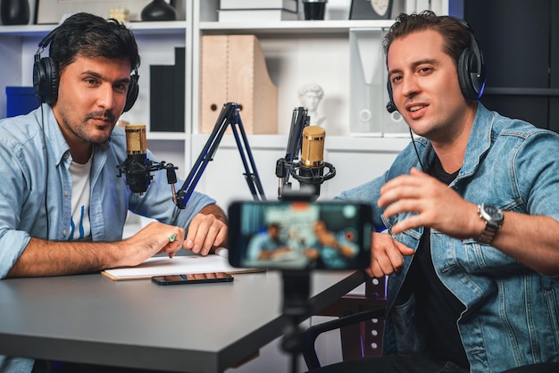 Foto anfitrión del canal de un locutor creativo que vive por teléfono inteligente en un podcast positivo con un invitado especial en la lista de guiones para aconsejar a los oyentes que lleven auriculares usando micrófono grabación de radio en el estudio sellable